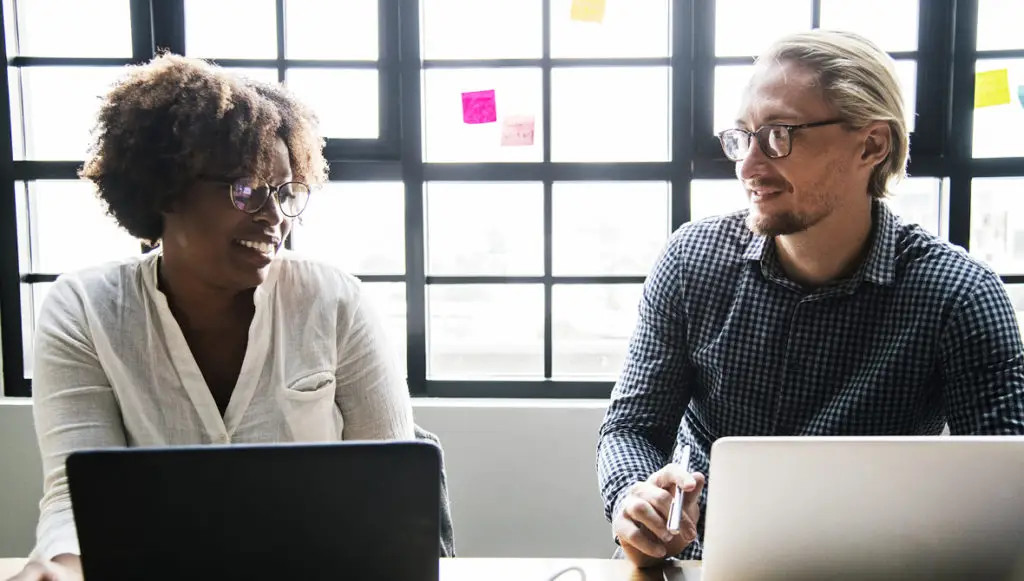Photo of a black femal professional and a white mal professional sitting side by side, chatting at a conference table with laptops in front of them, reseprenting two people discussing a theory of philanthropy