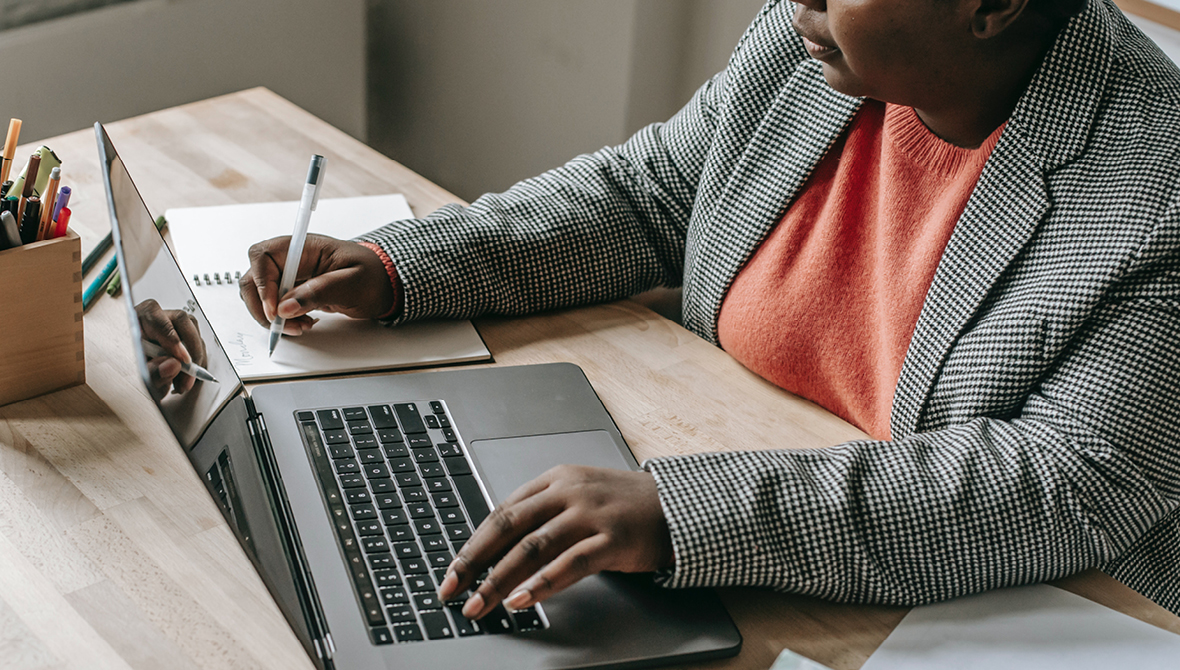 A Black woman wearing a checked-patterned blazer and a coral-colored blouse types on a laptop with a small notebook next to her.