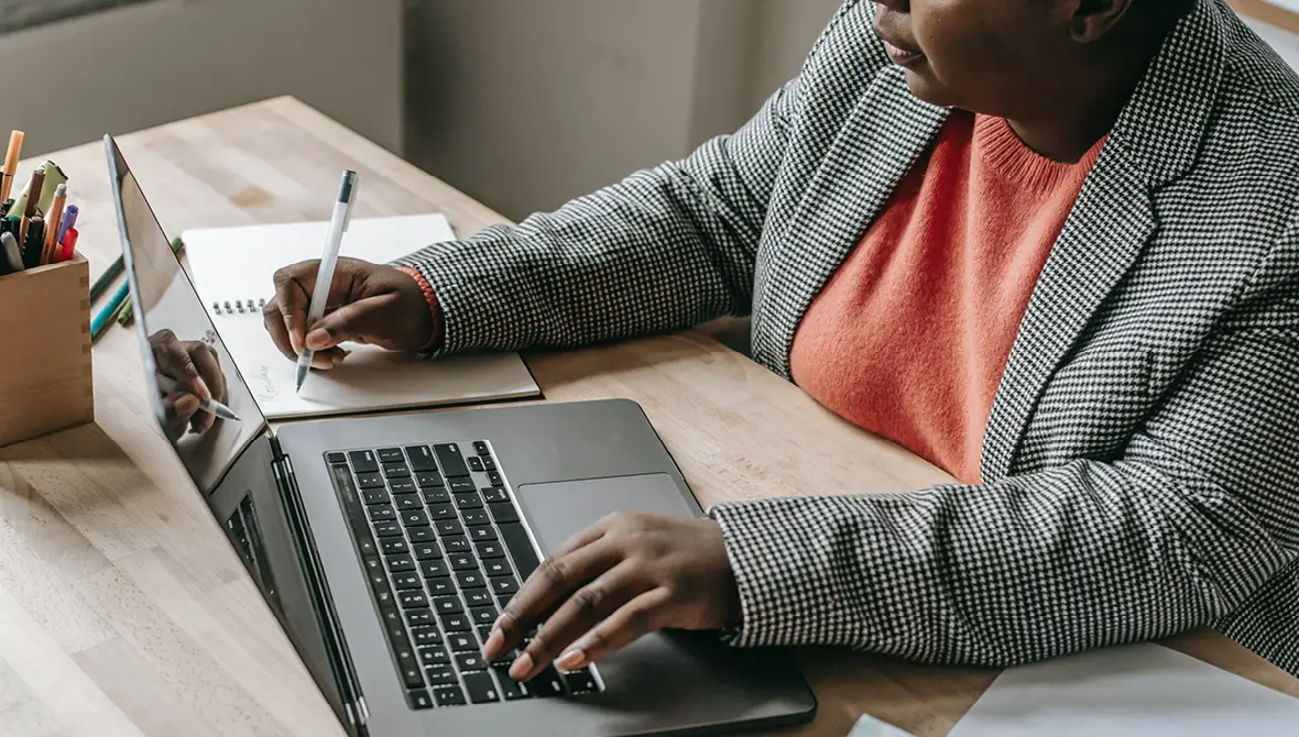 A Black woman wearing a checked-patterned blazer and a coral-colored blouse types on a laptop with a small notebook next to her.