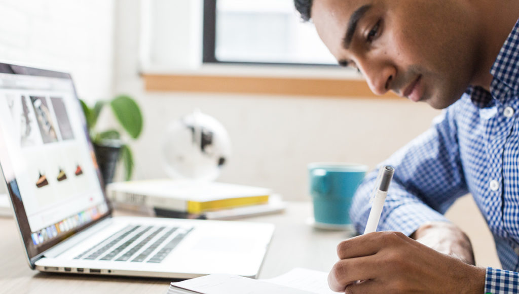 Photo of a man sitting in front of a laptop computer and writing in a notebook