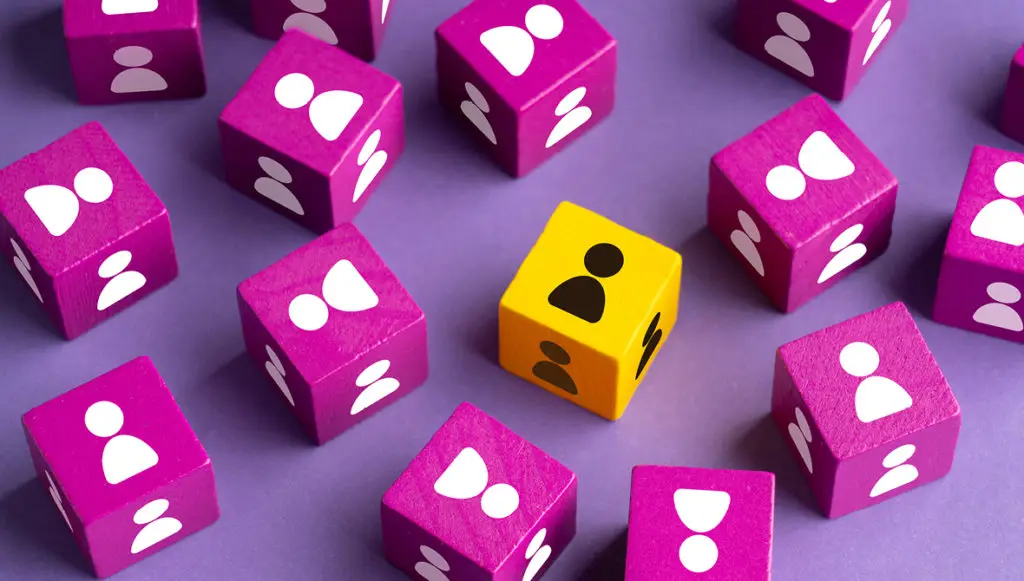 Photo of several bright purple blocks scattered on a table with a white human silhouette on each block. There is one yellow block that stands out, with a black human silhouette on it.