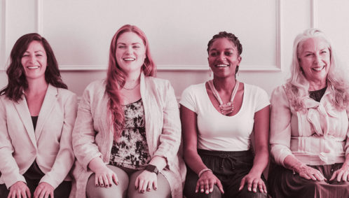 Photo of four, smiling adult women sitting next to each other in a row, each a different age, ethnicity, and body type.