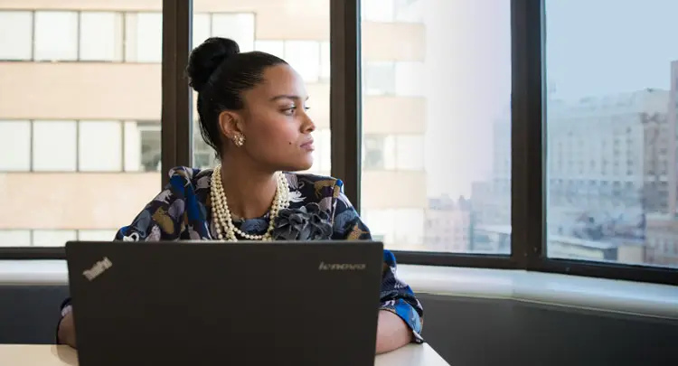Photo of a young, Black businesswoman sitting at a table in front of a laptop computer, looking off into the distance in thought (presumably about resilience and capacity building)