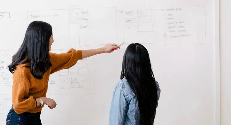 Photo of two women talking in front of a whiteboard with note written on it