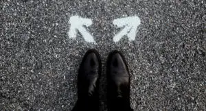 Photo of a man's feet wearing black dress shoes, standing on a road with two white arrows spray painted on the asphalt in front of his feet, pointing in opposite directions.