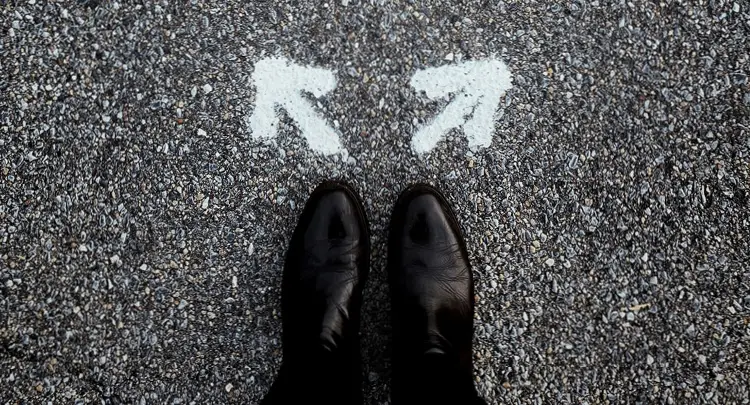 Photo of a man's feet wearing black dress shoes, standing on a road with two white arrows spray painted on the asphalt in front of his feet, pointing in opposite directions.