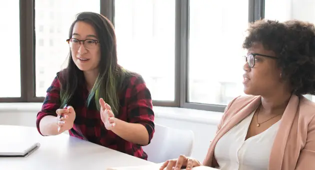 Photo of two woman sitting at a conference table talking to someone off camera