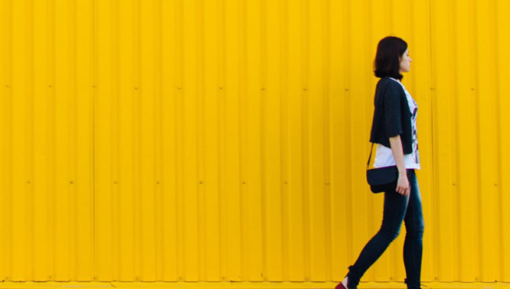 Photo of a professional young woman walking in front of large, bright yellow siding on a building