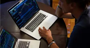 Photo of a man sitting at a table with two laptops, each showing charts of data.
