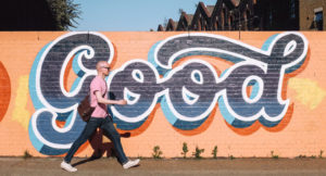 Photo of a man walking on a sidewalk in front of a brick wall with graffiti and the word "Good" painted on it