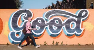 Photo of a man walking on a sidewalk in front of a brick wall with graffiti and the word "Good" painted on it