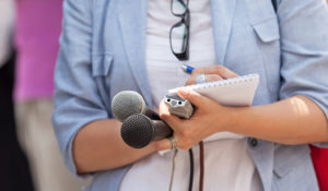 Photo of a journalist writing on a notepad while holding two microphones and a recording device.