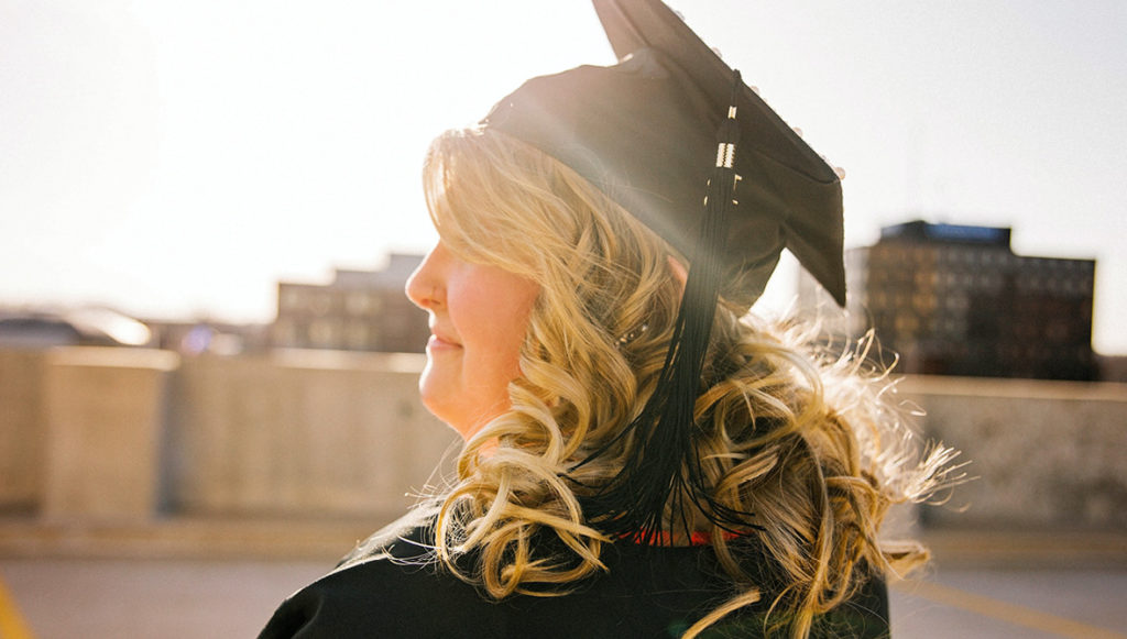 Photo of an adult woman wearing a black, academic mortarboard cap & gown, representing nontraditional alumni