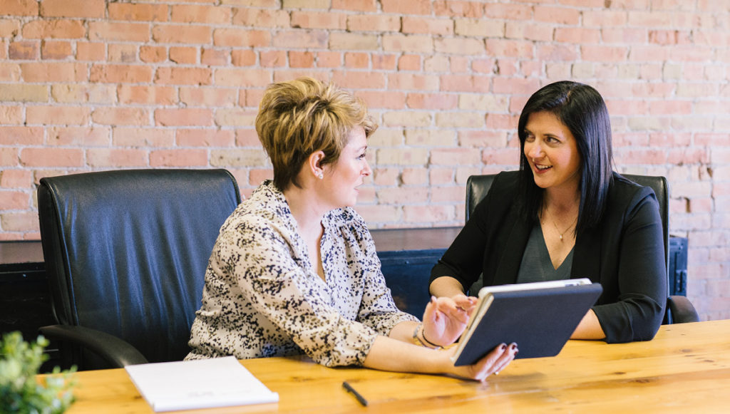 Two businesswomen sitting at a conference table, talking and looking at a laptop
