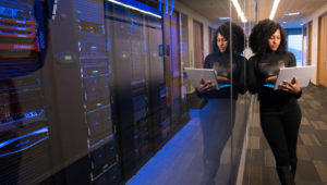 Photo of a Black woman standing next to a bank of servers and looking at a laptop