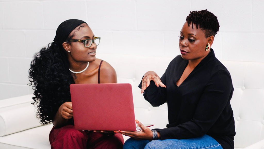 Photo of two Black women sitting on a couch, looking at a laptop and discussing a concern