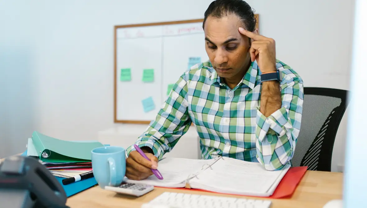 Man in a green checkered dress shirt sits at his desk looking concerned over papers in a binder in front of him