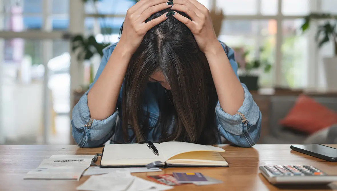 A woman with dark hair, sitting at a table surrounded by financial papers and a calculator, looks down, holding her head in her hands
