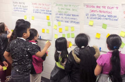 Photo of children gathered around large posters on a classroom wall with sticky notes on them. Photo courtesy of The Giving Square.