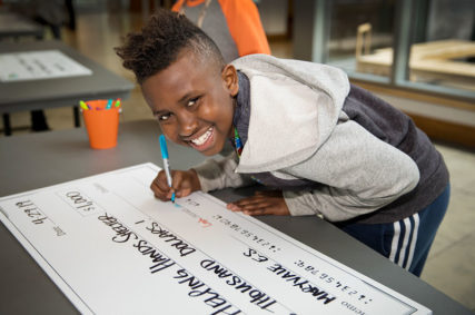 Photo of a young Black boy smiling and looking at the camera as he signs his name to a large novelty check representing a donation to a charity. Photo courtesy of The Giving Square.