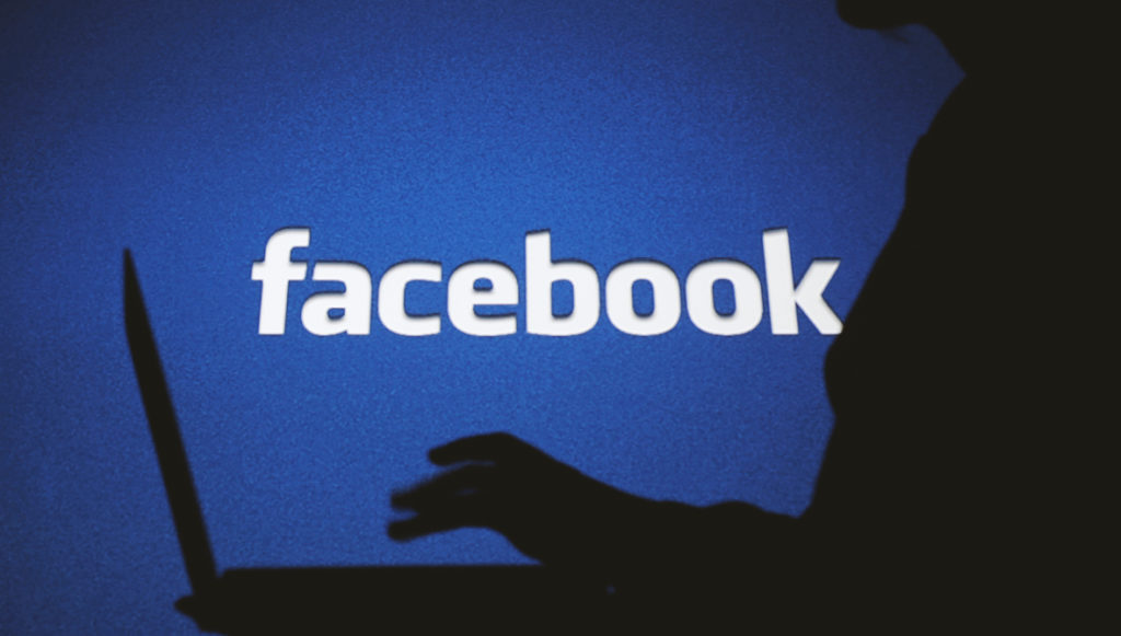 Photo: Dark silhouette of a woman typing on a computer in front of a backlit sign of the Facebook logo. Photo credit: Alexandra Popova