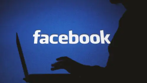 Photo: Dark silhouette of a woman typing on a computer in front of a backlit sign of the Facebook logo. Photo credit: Alexandra Popova