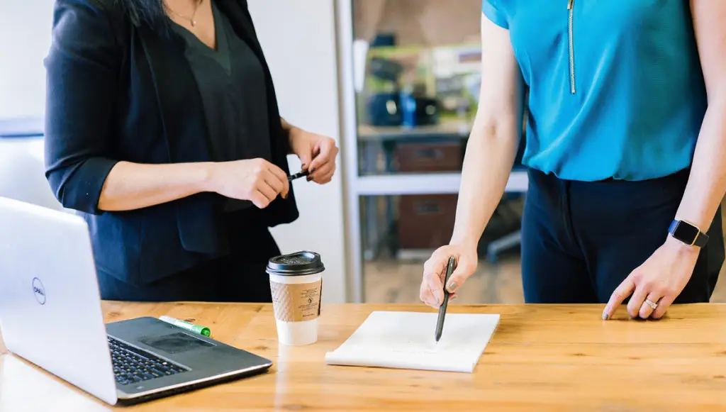Photo of two business women standing behind a conference table pointing to a paper and a laptop on the table. (photo represents communicating corporate philanthropy to business partners)ropy