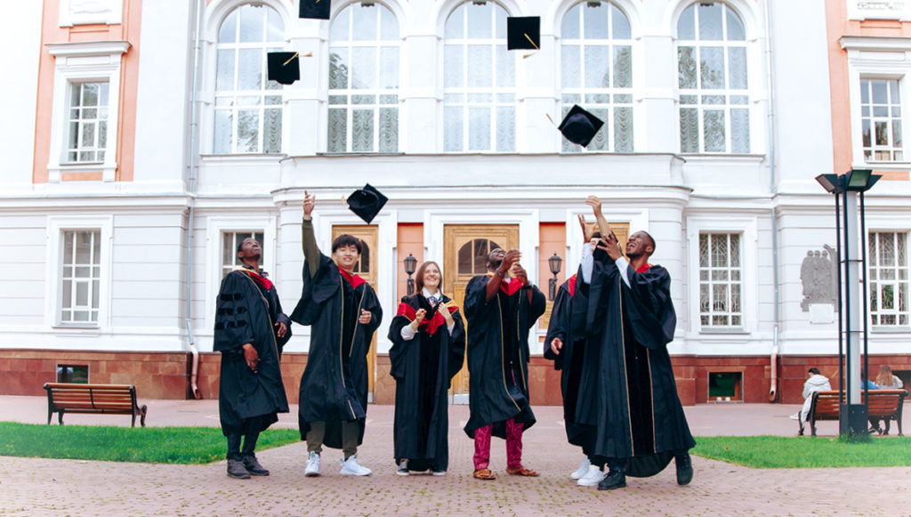 Six graduates stand outside of a college building in their black graduation robes, throwing their caps into the air, representing post-secondary education