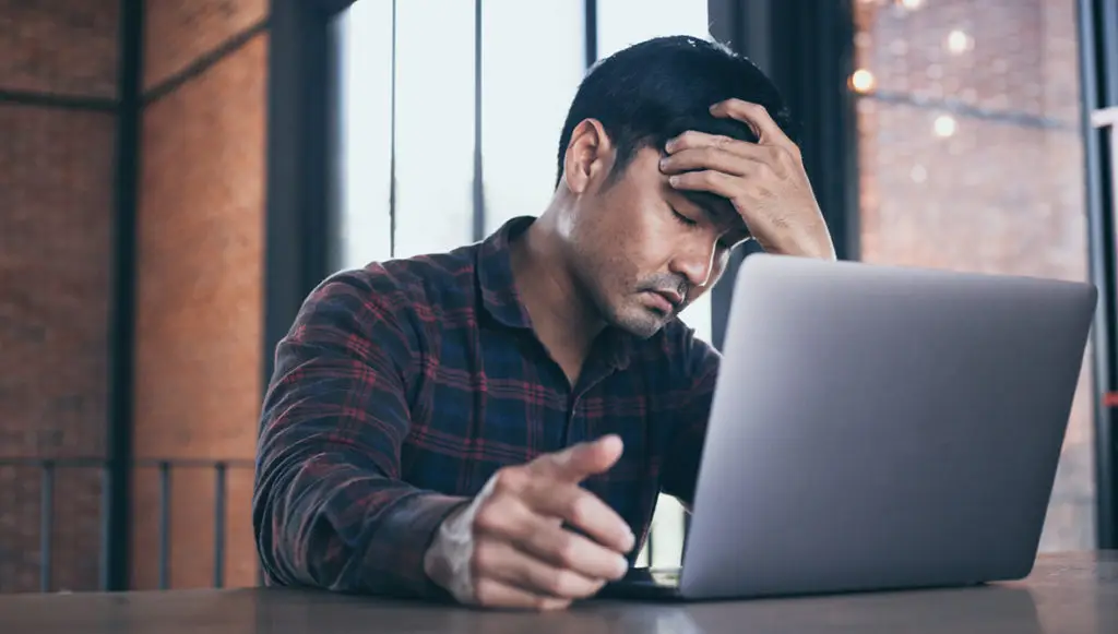 Photo of an Asian man sitting in front of a laptop with his hand on his forehead as if frustrated or stressed (representing the feeling of burnout)