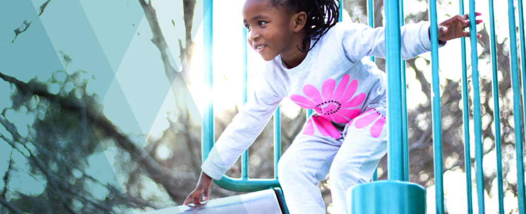 A young, smiling Black girl wearing braids and a gray sweatshirt with a bright pink flower on the front climbs to the top of a playground slide