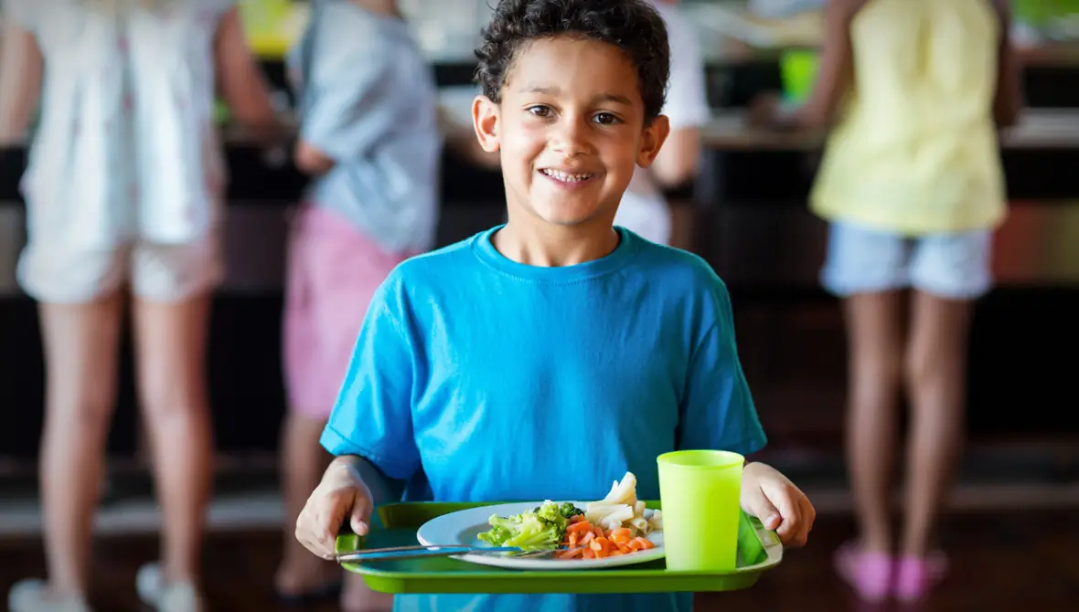 A young boy in a blue t-shirt holds a green school lunch tray with a plate of fruits and vegetables on it, representing health equity