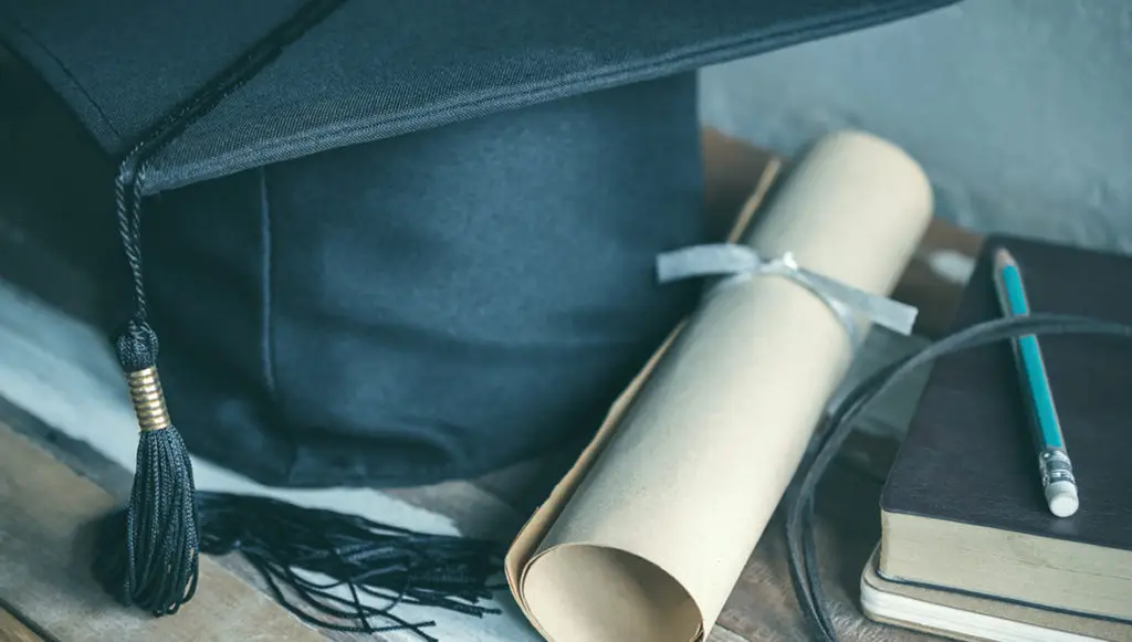 Photo of a black graduation cap next to a rolled-up diploma tied with a white ribbon and a stack of research books