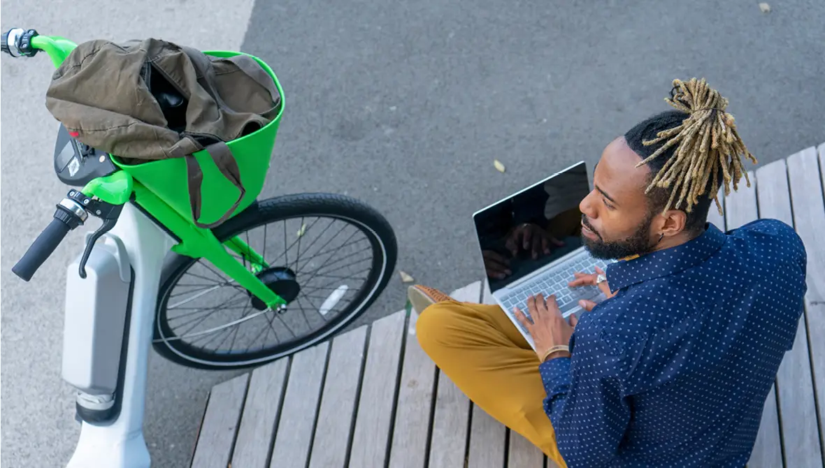 Photo of a young Black man wearing a blue button-down shirt and khaki pants sitting on a bench, using a laptop, next to a bike from a bike-share program (representing the sharing economy)