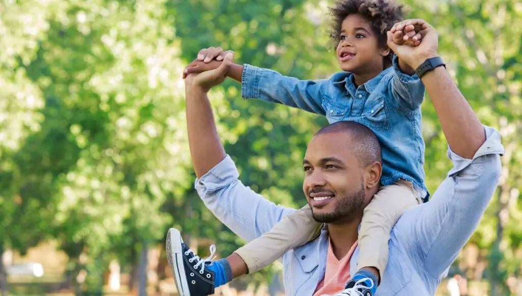 A Black father carries his happy, young son on his shoulders in a public park