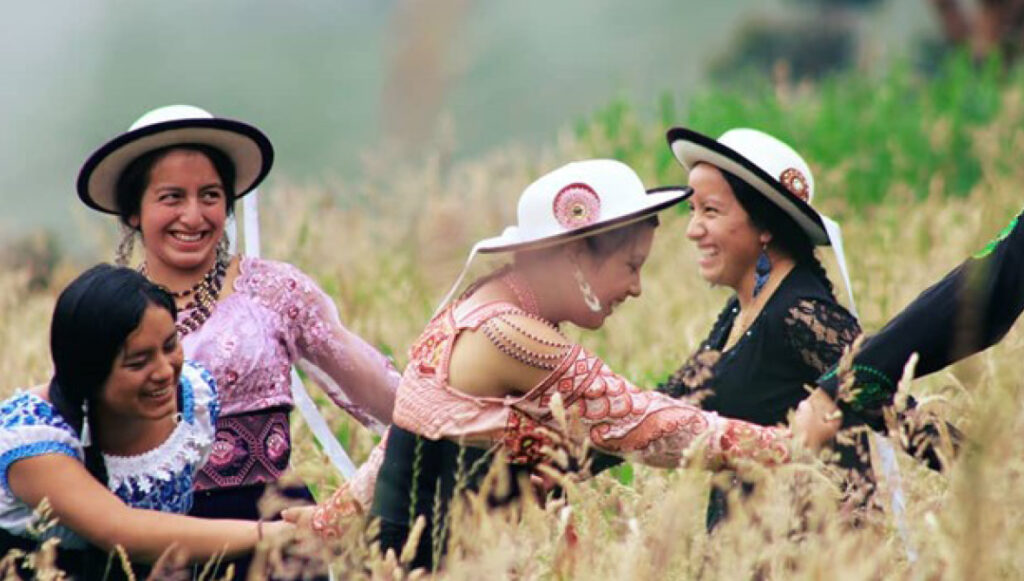Five smiling, Indigenous women hold hands, dancing in a circle in a field.