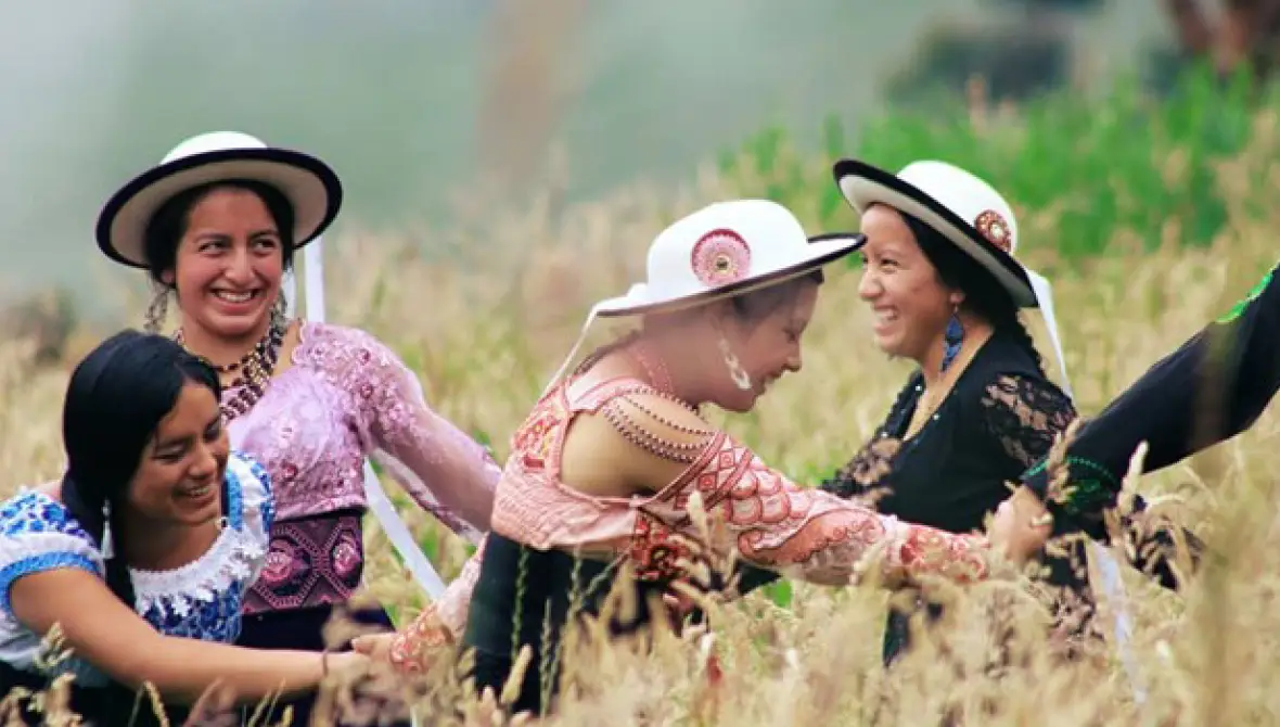 Five smiling, Indigenous women hold hands, dancing in a circle in a field.