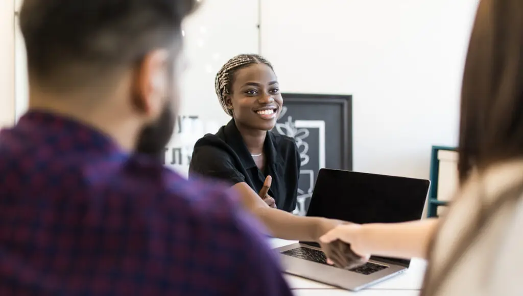 A Black woman wearing braids and a black blazer smiles and shakes the hand of her colleague, symbolizing a sense of belonging