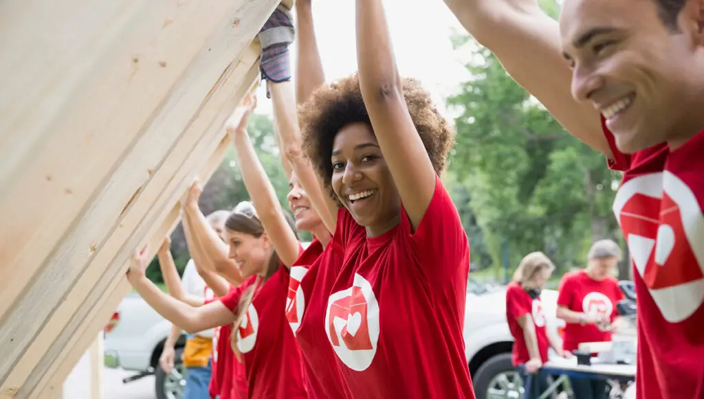 A group of volunteers wearing red shirts work together to build housing for those in need. In the center of the photo is a smiling young Black woman.