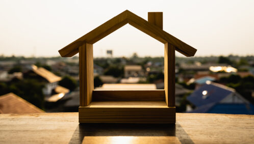 Photo of a small, wooden house figurine on a balcony overlooking a neighborhood of affordable housing