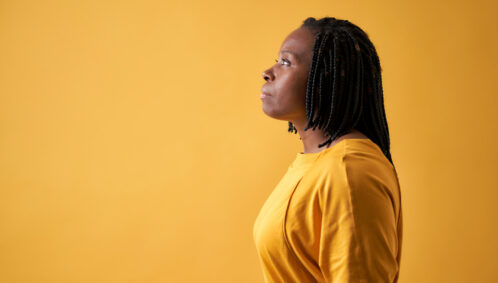 A Black woman wearing braids and a yellow blouse looks off to the distance in serious thought (as if to represent facing a glass cliff)