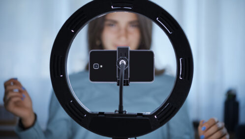 A young woman (representing a social media "influencer") stands in front of a ring light to record a video on her phone