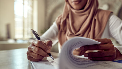A woman wearing a headscarf writes on a stack of papers in front of her on a table