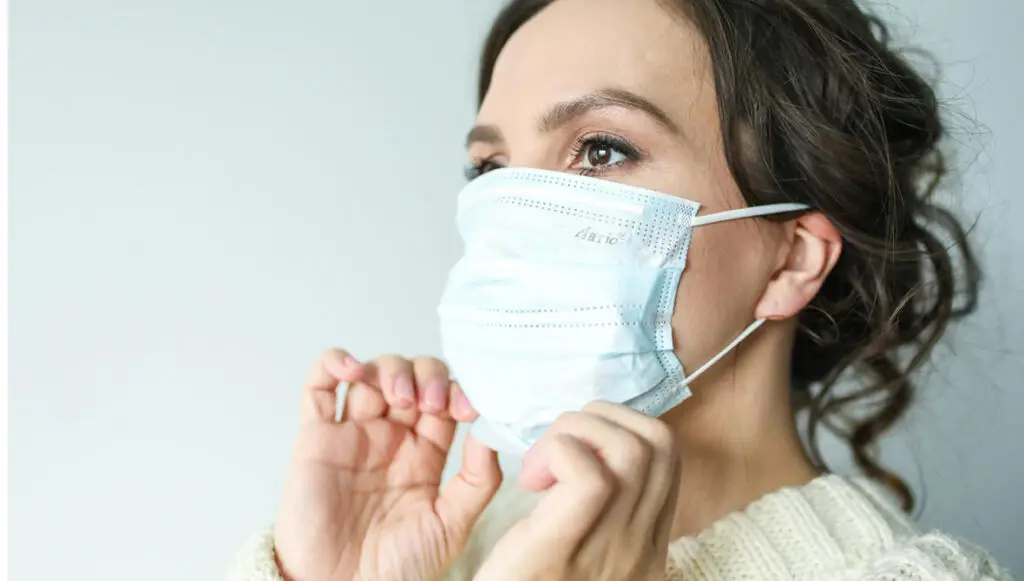 A woman with dark hair and brown eyes pulls a mask over her mouth and nose