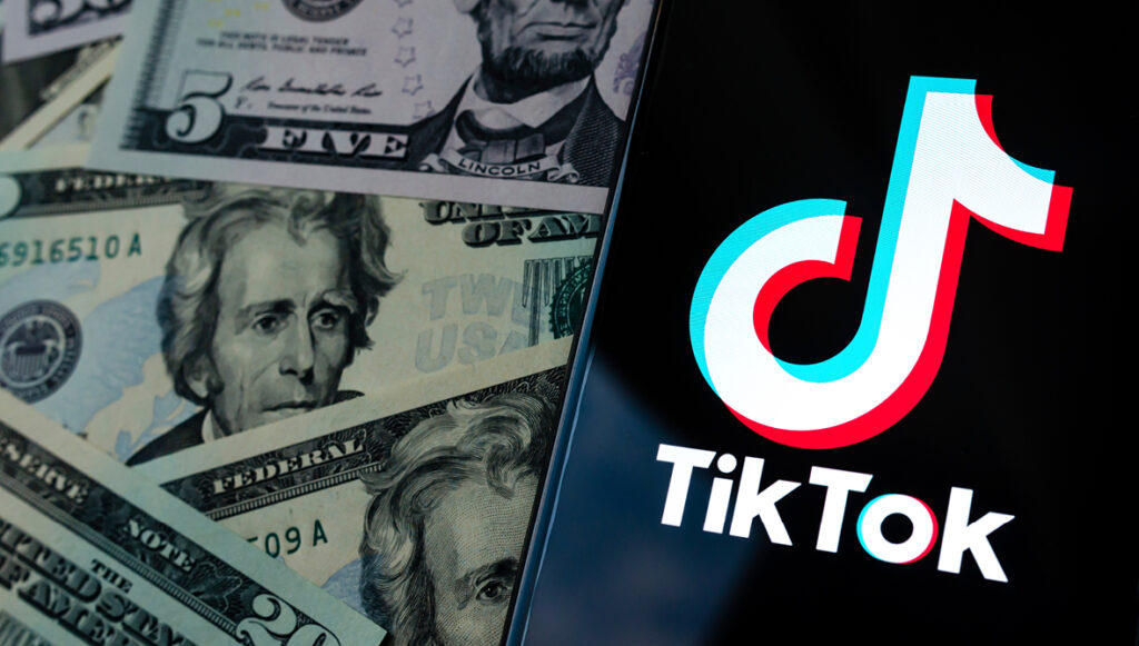 A mobile phone with the TikTok logo laying on a pile of dollar bills of different denominations