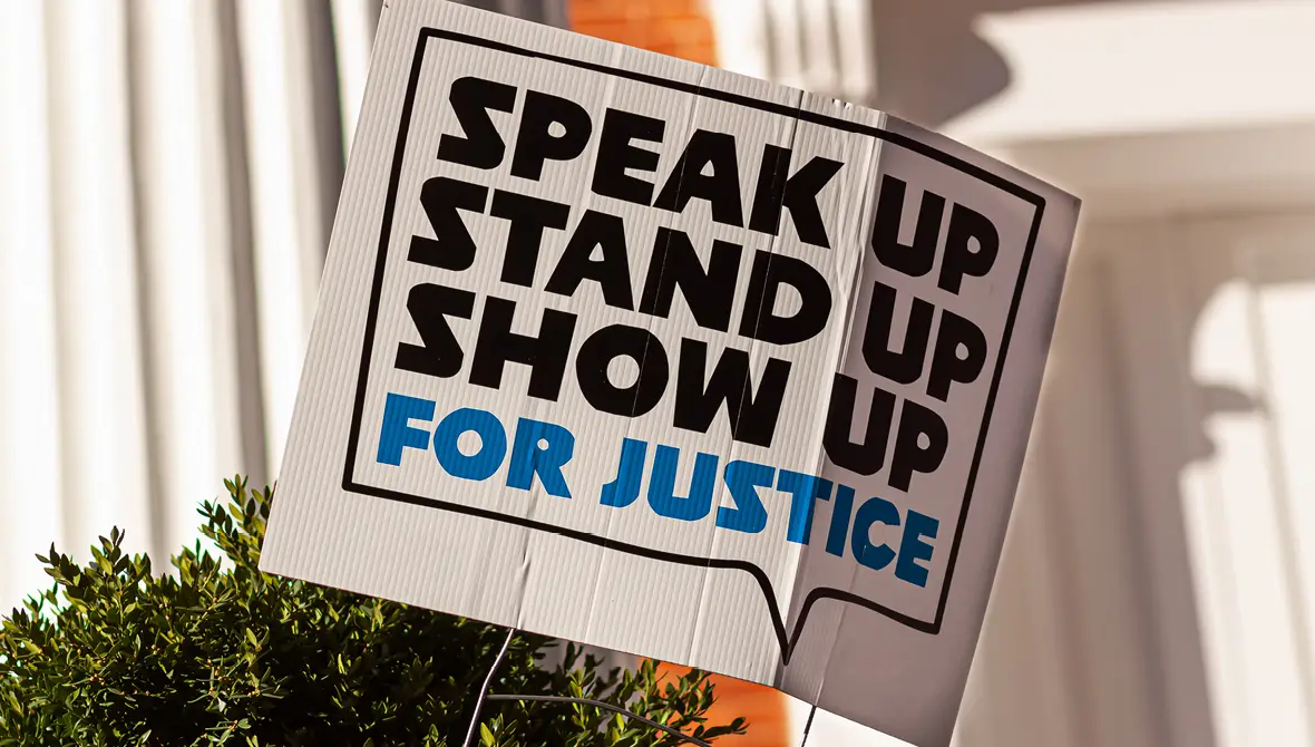 A yard sign reads "Speak Up. Stand Up. Show Up. For Justice." in large block-print letters.