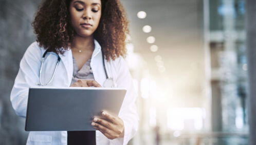 A young, Black woman wearing a white lab coat types on a tablet