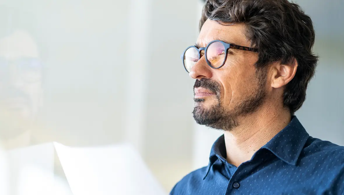 A man wearing dark-framed glasses looks off into the distance in thought