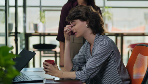 A woman sits at a desk with her eyes closed and her hand on her head in frustration