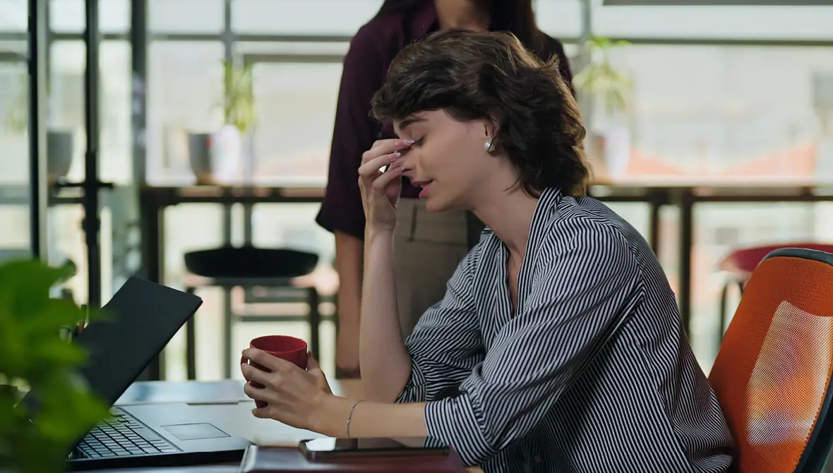 A woman sits at a desk with her eyes closed and her hand on her head in frustration