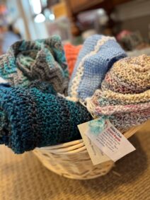 Basket of colorful knitted shawls and caps with a tag indicating the items were knit by volunteers at Gilda's Club.